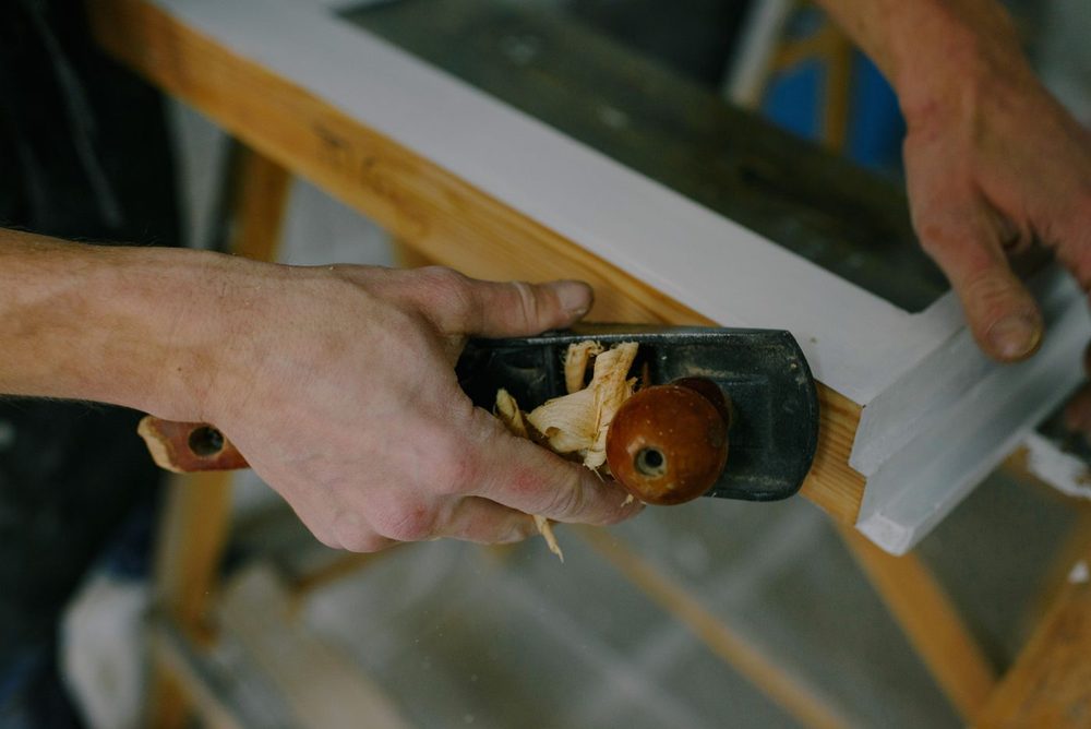 Carpenter running a hand plane along a painted window frame, shavings curling off the blade
