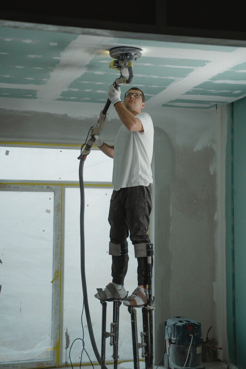 Drywaller on stilts sanding a taped ceiling with a dust-extraction sander